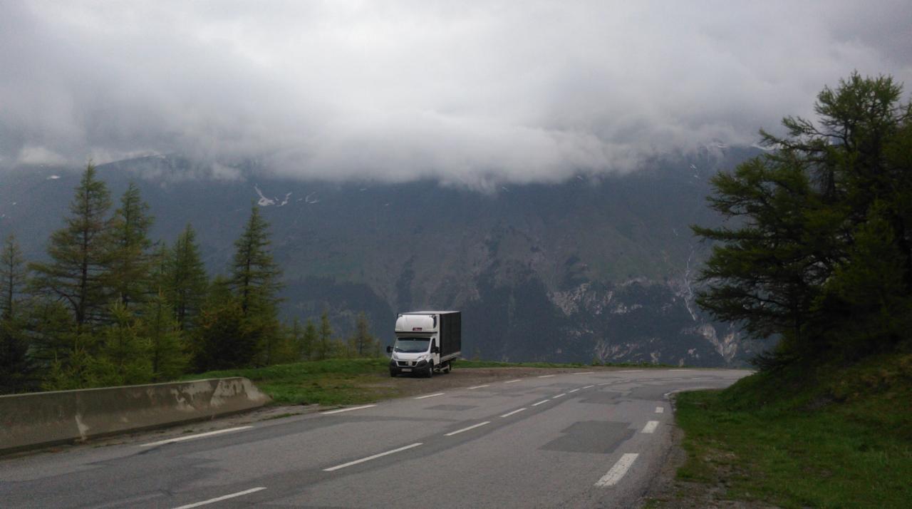 Samochód dostawczy zaparkowany na poboczu górskiej drogi z widokiem na zalesione zbocza i nisko wiszące chmury.