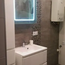 Nowoczesna łazienka z podświetlanym lustrem, umywalką z szafką w kolorze białym, grzejnikiem gazowym i dekoracyjną mozaiką w pionowym pasie na szarej ścianie.