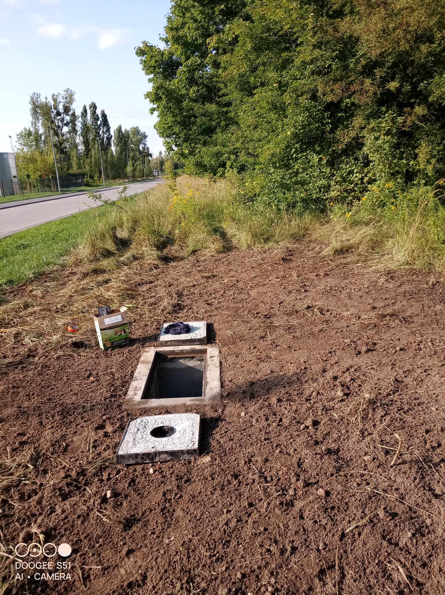 Otwarty właz betonowy w ziemi, obok karton i pokrywa, teren przygotowany pod instalację, widoczne trawy i drzewa w tle.