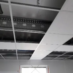 Montaż sufitu podwieszanego kasetonowego z widoczną konstrukcją nośną i instalacjami w przestrzeni międzystropowej, ujęcie z perspektywy pomieszczenia z oknem.