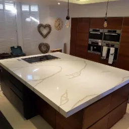 Nowoczesna wyspa kuchenna z białym blatem o marmurowym wzorze i wbudowaną płytą grzewczą, otoczona drewnianymi szafkami i sprzętem AGD, oświetlona wiszącymi lampami edisonowskimi.