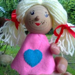 Amadeusz-Alina Krystel - Ręcznie robiona lalka z drewnianą główką, wełnianymi włosami związanymi czerwonymi wstążkami, ubrana w różową sukienkę z niebieskim sercem, trzymana w dłoni na tle zieleni.