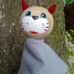 Amadeusz-Alina Krystel - Ręcznie robiona kukiełka kota z drewnianą główką, czerwonymi uszami i szarym filcowym ubrankiem, trzymana na tle kory drzewa.