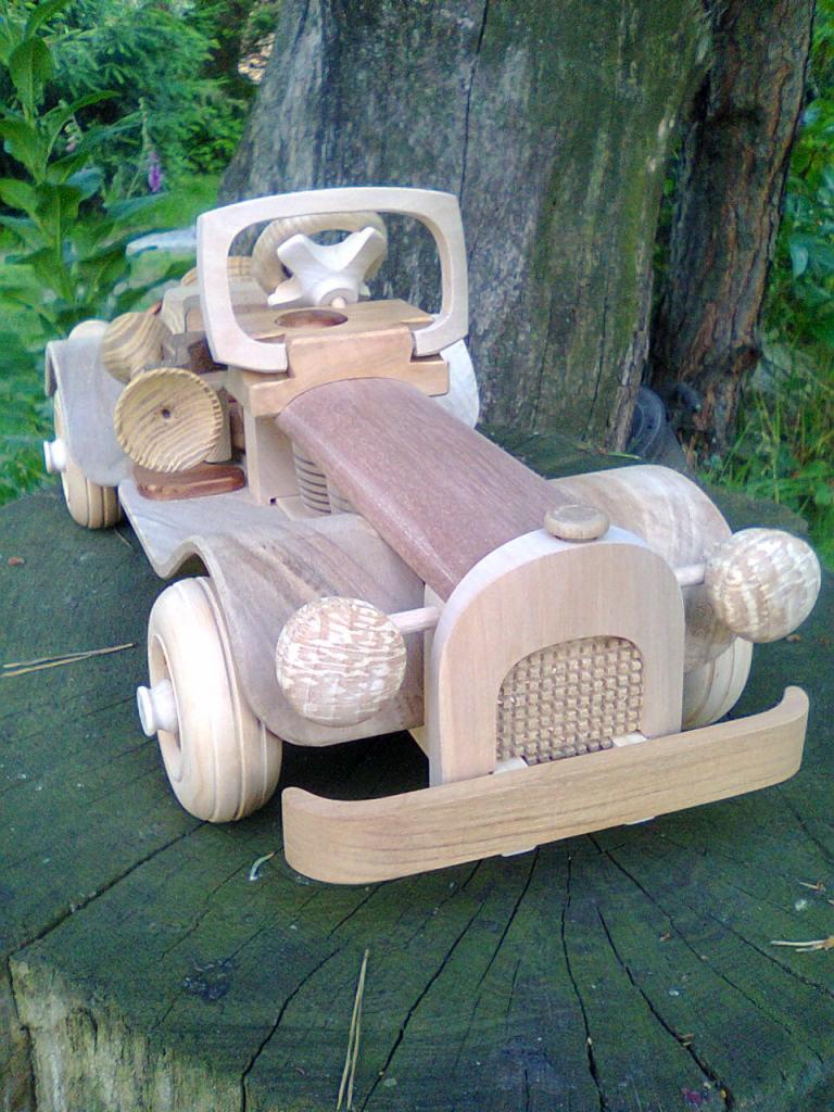 Ręcznie wykonany, drewniany model samochodu retro na pniu drzewa, z widocznymi detalami snycerskimi i fakturą drewna.