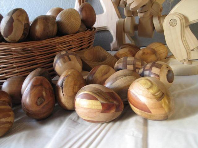 Ręcznie wykonane, drewniane jajka dekoracyjne w koszyku wiklinowym i obok, niektóre z intarsjami i wzorami z różnych gatunków drewna, na białym tle.