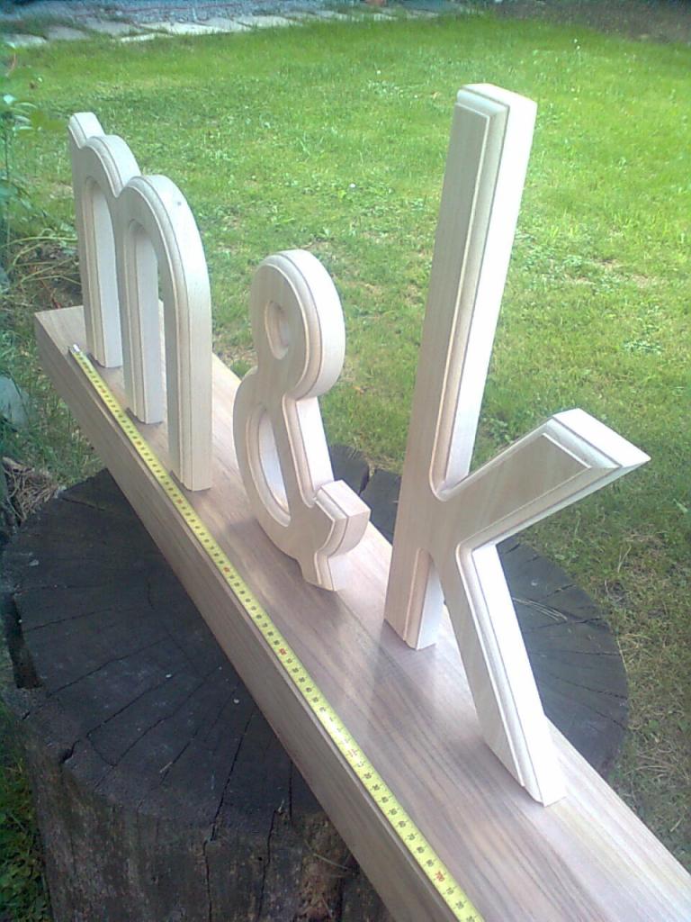 Surowe, drewniane litery 'm&k' ułożone na desce z miarką, stojące na pniu drzewa na tle trawy.