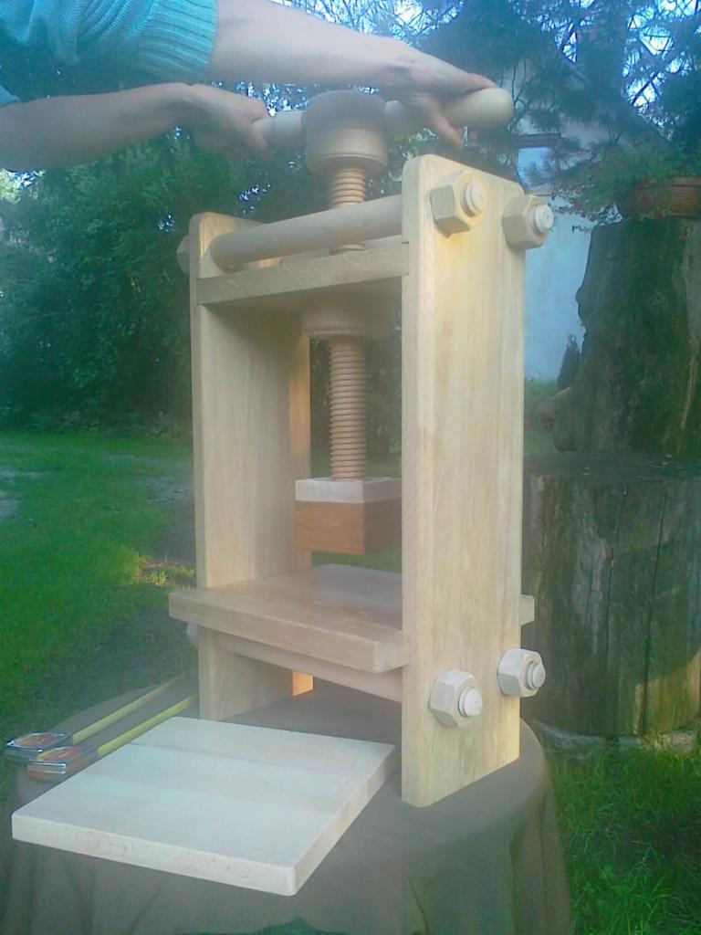 Drewniana prasa śrubowa obsługiwana ręcznie, umieszczona na stole w ogrodzie, z widocznymi narzędziami obok.