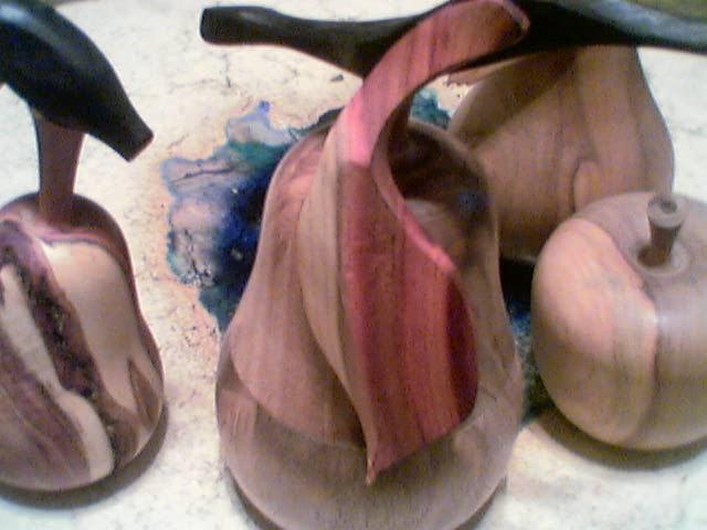 Ręcznie rzeźbione, drewniane figurki owoców: gruszka z czarną szypułką, otwarta gruszka z czerwonym wnętrzem oraz jabłko z brązową szypułką, na tle z plamami farby.