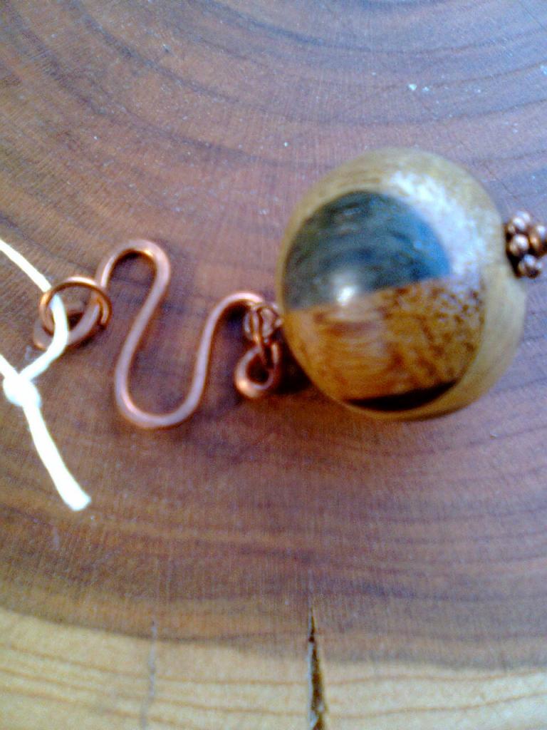 Drewniana kula z miedzianymi elementami i białym sznurkiem na tle drewnianego przekroju pnia z widocznymi słojami.