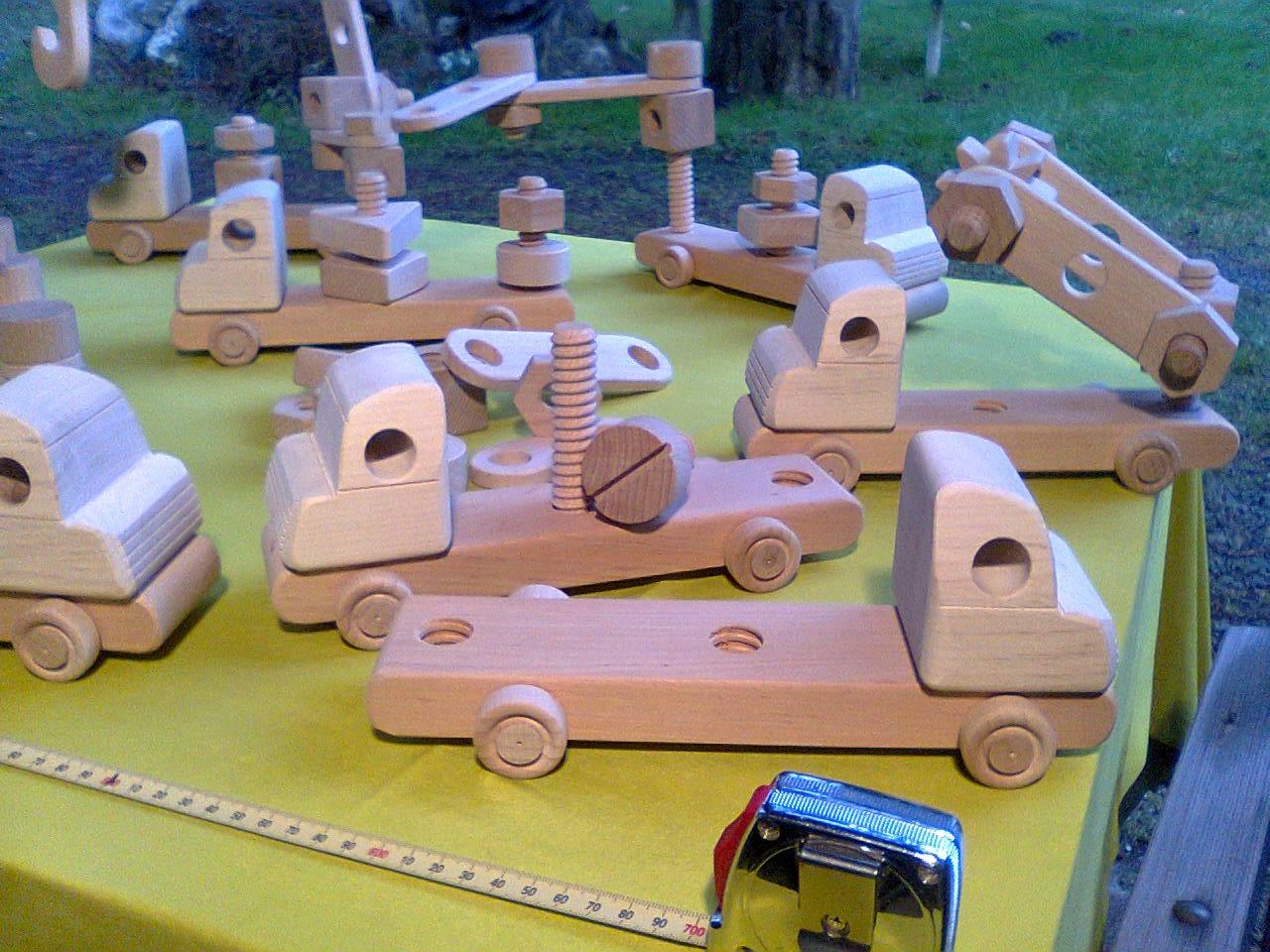 Ręcznie robione drewniane zabawki: ciężarówki i dźwig na żółtym tle, z miarką zwijaną w rogu, sugerujące pomiar lub prezentację rozmiaru.