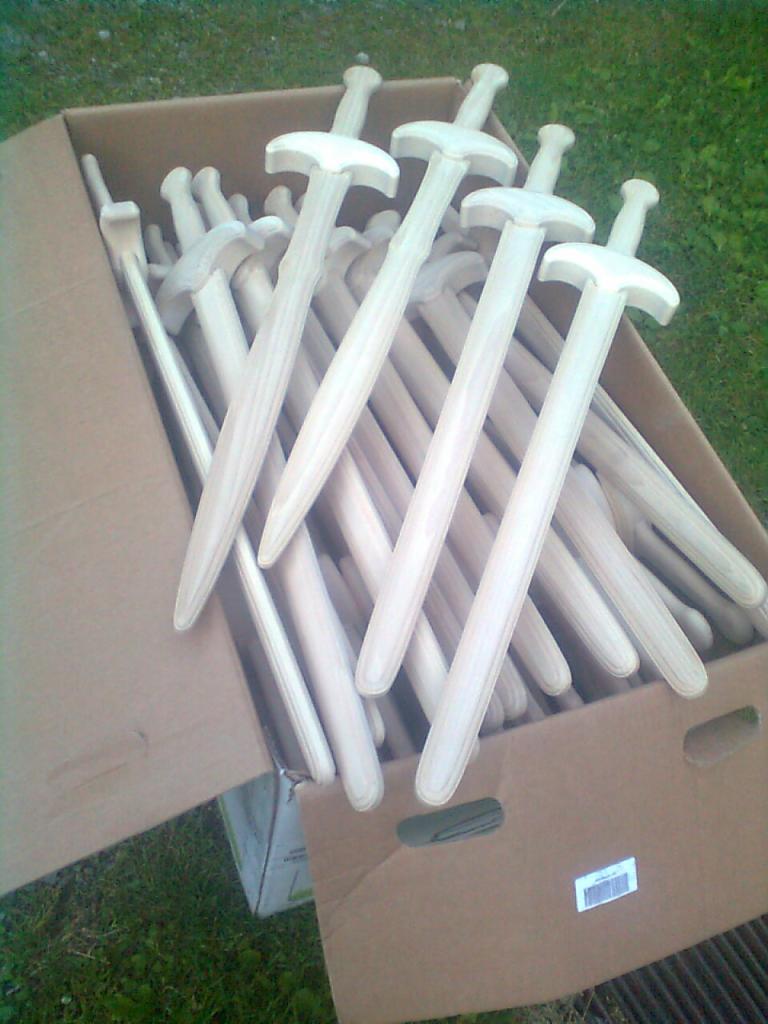 Kartonowe pudło wypełnione serią drewnianych mieczy z jasnego drewna, ułożonych warstwami, widoczne na zewnątrz na tle trawy.
