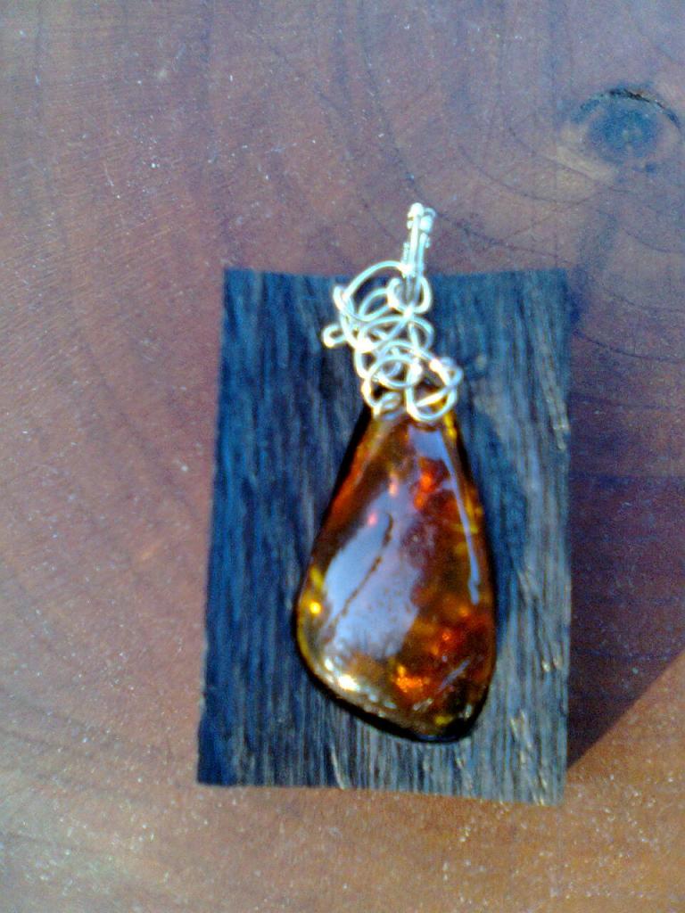 Bursztynowy wisiorek w kształcie łzy, opleciony srebrnym drutem, prezentowany na kawałku ciemnego drewna na tle drewnianej powierzchni.