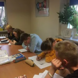 Grupa uczniów skupiona na pisaniu przy stole w jasnej sali lekcyjnej, widoczny plakat z napisem 'Let's Speak' i rośliny na parapecie.