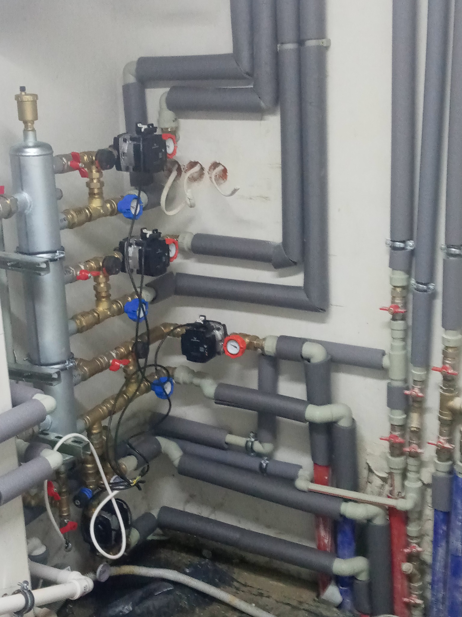 Złożona instalacja grzewcza z rurami w izolacji termicznej, zaworami i pompami obiegowymi, zamontowana przy białej ścianie. Widoczne elementy miedziane i plastikowe.