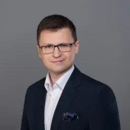 Portret prawnika w okularach, ubrany w ciemny garnitur z wzorzystą poszetką, uśmiechający się na szarym tle.