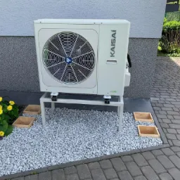 Zewnętrzna jednostka klimatyzacji marki Kaisai, zamontowana na metalowym stelażu przy ścianie budynku, otoczona dekoracyjnym żwirem i kwitnącymi żółtymi kwiatami.