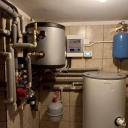 A-Z INSTALACJE Sp. z o.o. - Kotłownia z dwoma zbiornikami, pompą obiegową i osprzętem, widoczna instalacja rur z izolacją, płytki ceramiczne na ścianach i podłodze.