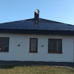 Jednorodzinny dom z ciemną dachówką i panelami słonecznymi na dachu, z anteną satelitarną na kominie, widok z boku na ogród.