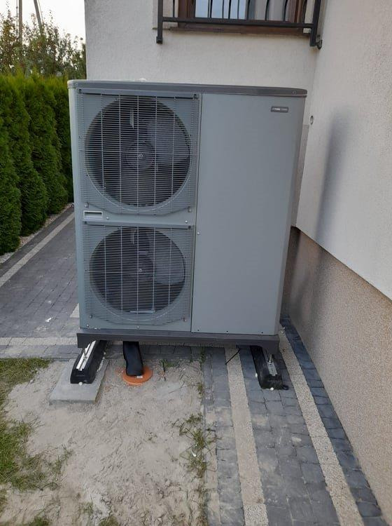 Szara pompa ciepła typu powietrze-woda z dwoma wentylatorami, zamontowana na betonowych podstawach obok budynku z widoczną elewacją i fragmentem okna.