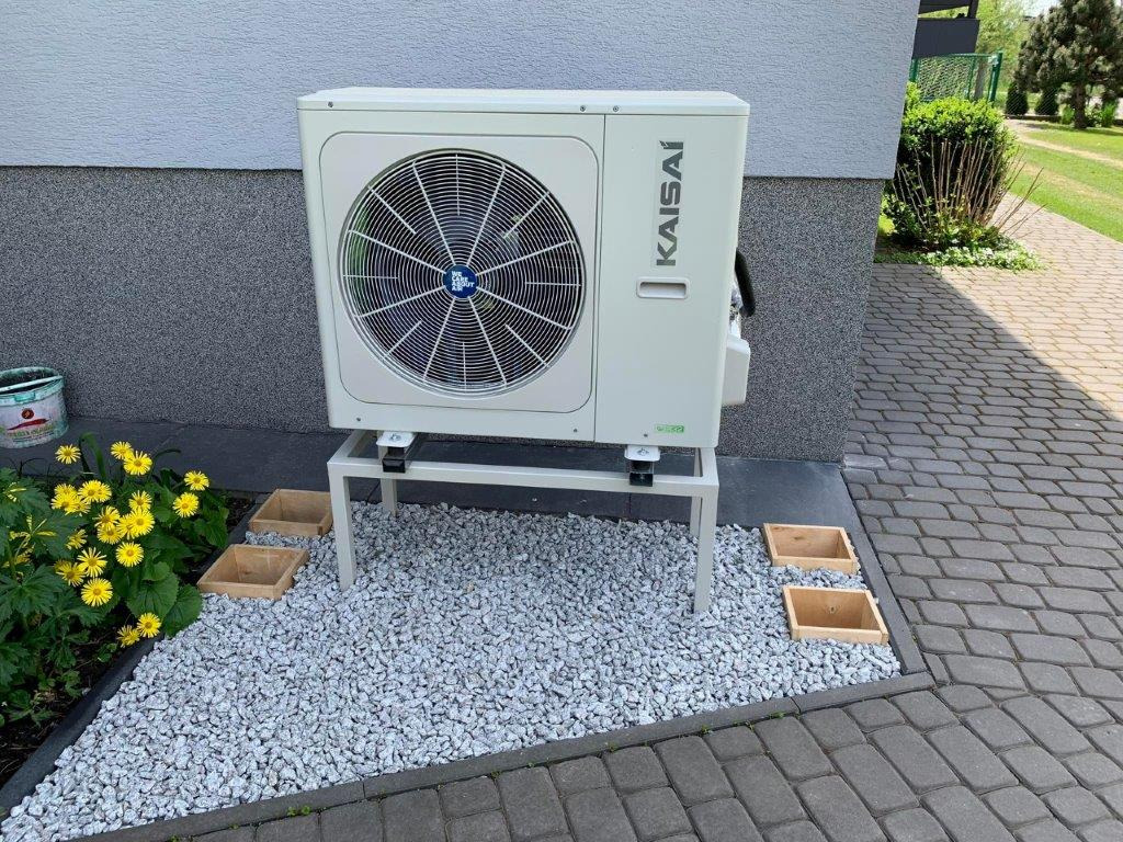 Zewnętrzna jednostka klimatyzacji marki Kaisai, zamontowana na metalowym stelażu przy ścianie budynku, otoczona dekoracyjnym żwirem i kwitnącymi żółtymi kwiatami.