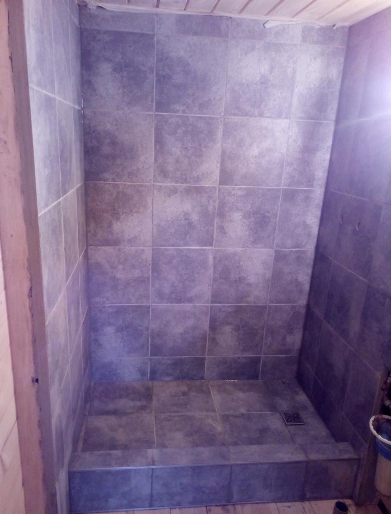 Wykończony brodzik z szarych, kwadratowych płytek ceramicznych, z widocznym odpływem liniowym, drewnianą boazerią i fragmentem wiadra w rogu.