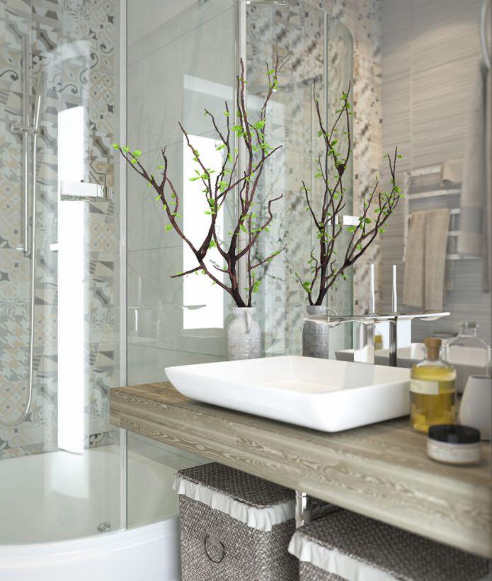 Nowoczesna łazienka z umywalką nablatową o kwadratowym kształcie, drewnianym blatem, dekoracyjnymi gałązkami w wazonach, szklaną kabiną prysznicową i koszami z wikliny pod blatem.