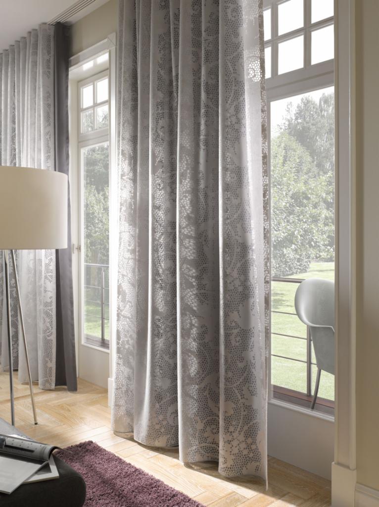 Delikatne, wzorzyste zasłony w odcieniach szarości i bieli zawieszone w jasnym salonie z widokiem na zieleń za oknem.