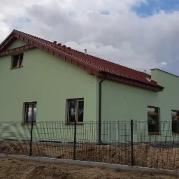 Świeżo otynkowany, parterowy dom z czerwonym dachem, widoczny zza metalowego ogrodzenia. Na podwórku stoi traktor, a w tle widać inny, żółty budynek.