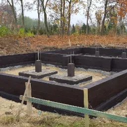 Wykopane i zabezpieczone fundamenty pod budynek, z zalanymi betonem słupami i wystającymi prętami zbrojeniowymi, na tle jesiennego krajobrazu z drzewami w odcieniach żółci i brązu.