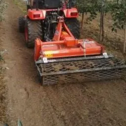 Czerwony traktor z glebogryzarką przygotowuje ziemię pod sadzenie, widok z tyłu, ziemia lekko wilgotna, sadzonki drzew w tle.