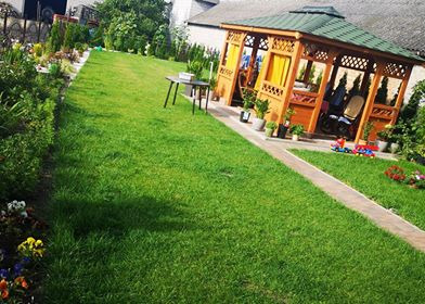 Zadbany ogród z równo przystrzyżonym trawnikiem, drewnianą altaną z żółtymi zasłonami, rabatami kwiatowymi i ścieżką z kostki.