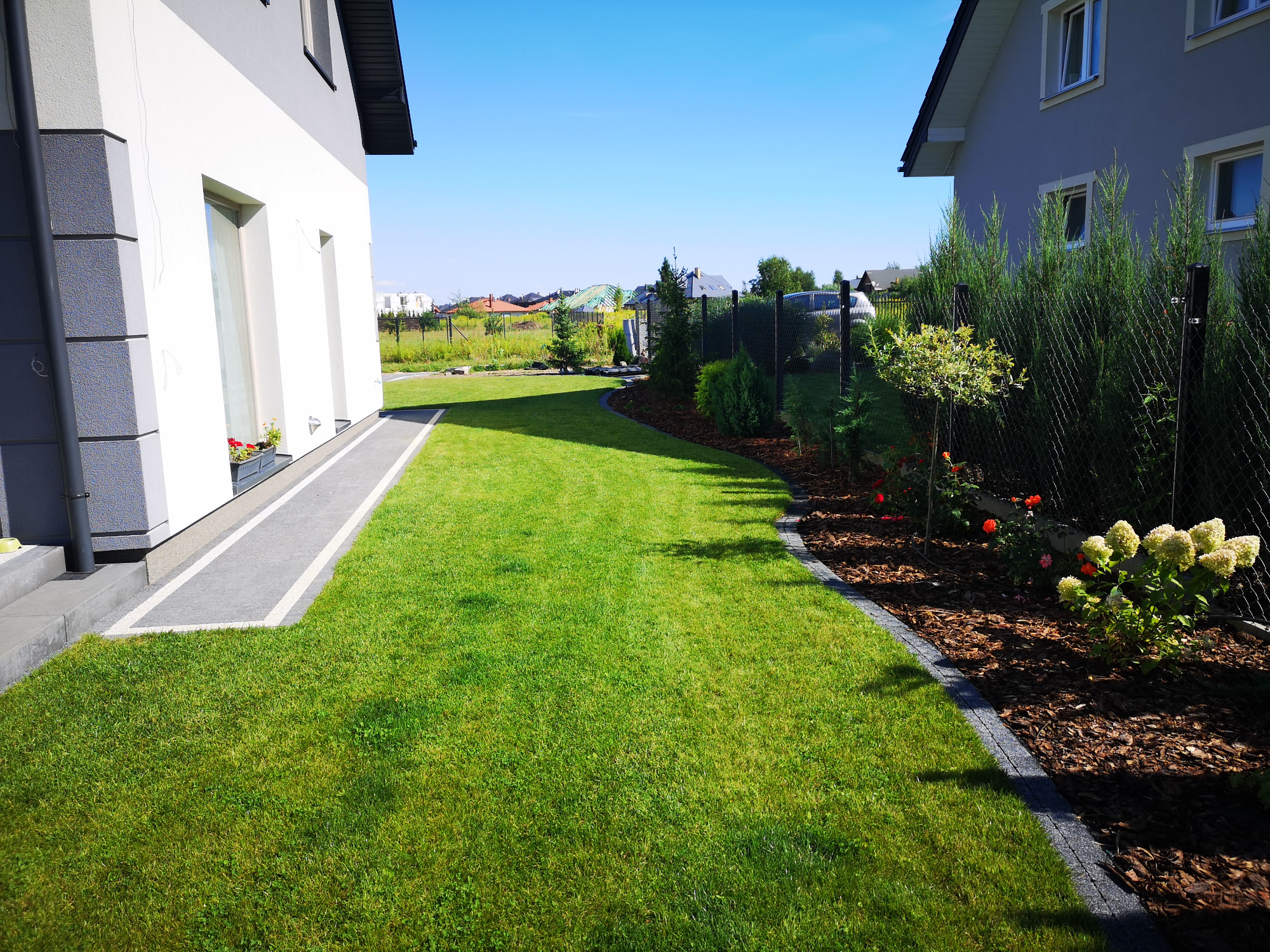 Zadbane podwórko z równo przystrzyżonym trawnikiem między dwoma nowoczesnymi domami, widoczne rabaty kwiatowe i obrzeża z kory.
