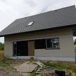 Niedokończony dom szkieletowy z ciemnoszarą dachówką, widoczne okno dachowe i nieotynkowana podstawa, teren zarośnięty trawą.