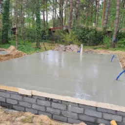 Wylany, mokry beton na fundamencie z bloczków betonowych w trakcie budowy domu, z widocznymi niebieskimi rurami wystającymi z betonu i lasem w tle.