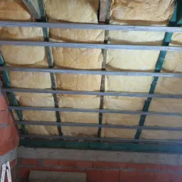 Ocieplenie wełną mineralną poddasza w trakcie prac w Łebczu, widoczne metalowe profile i ceglana ściana boczna.