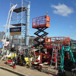 Szeroki kadr prezentujący różnorodny sprzęt budowlany na zewnątrz, w tym rusztowanie z banerem Arsenal, podnośnik nożycowy Snorkel GTJZ2088ES, minikoparka i przecinarki do betonu.