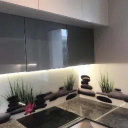 Szare szafki kuchenne z podświetleniem LED i panelem dekoracyjnym z motywem kamieni zen, trawy i różowego kwiatu lotosu nad płytą indukcyjną.