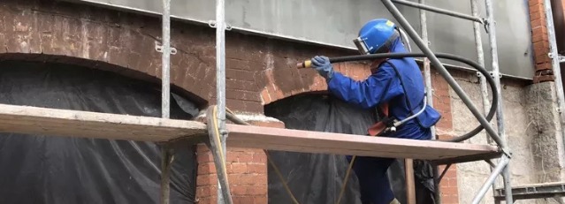 Pracownik w niebieskim kombinezonie i masce ochronnej piaskuje ceglaną elewację budynku z rusztowania, widoczne fragmenty oczyszczonej powierzchni i zabezpieczone okno.