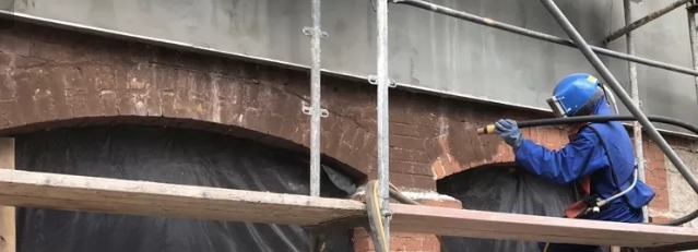 Pracownik w kombinezonie ochronnym, na rusztowaniu, czyści ceglany łuk na budynku za pomocą strumienia ściernego.