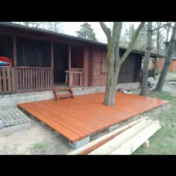 Świeżo pomalowany, brązowy taras z drewnianych desek, częściowo otaczający pień drzewa, z niskimi schodkami prowadzącymi do wejścia do drewnianego domku letniskowego.