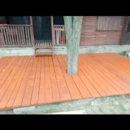 Nowy, pomarańczowy drewniany taras zbudowany wokół drzewa, z widocznym wejściem do drewnianego domu na podwyższeniu.