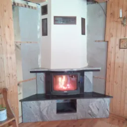 Kominek z czarnym obramowaniem i płomieniami w palenisku, obudowany szarą płytą kamienną, widoczne elementy drewnianej boazerii i przygotowania do wykończenia ściany