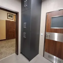 Wysoka, grafitowa szafa z czterema drzwiami i srebrnymi uchwytami, umieszczona w korytarzu obok drewnianych drzwi z metalowymi akcentami i małym okienkiem.