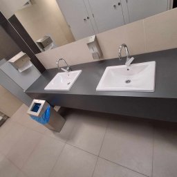 Master Furniture - Dwie białe umywalki nablatowe z chromowanymi bateriami na szarym blacie w publicznej toalecie, z dozownikami mydła i ręczników papierowych na ścianie.