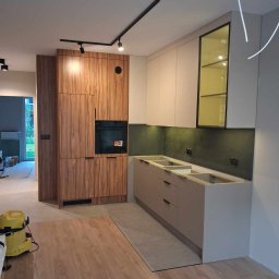 Master Furniture - Montaż kuchni na wymiar w trakcie: drewnopodobna zabudowa z piekarnikiem, szare szafki dolne bez blatów, zielony panel ścienny, czarne oświetlenie szynowe, żółty odkurzacz na podłodze.