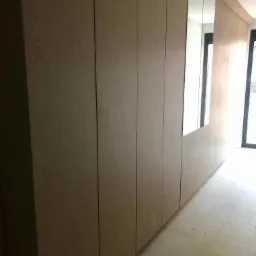 Długa, zabudowana szafa z jasnego drewna z wbudowanym lustrem, ciągnąca się wzdłuż korytarza z jasną podłogą i oknem w tle.