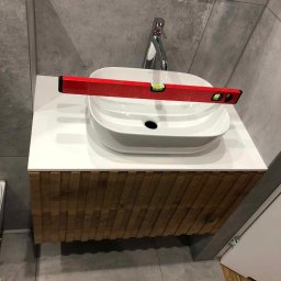 Master Furniture - Biała, owalna umywalka nablatowa z baterią, ustawiona na szafce z drewnianymi lamelami; poziomica spoczywa na krawędzi umywalki, sprawdzając jej wypoziomowanie na blacie; ściana wykończona szarą...