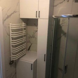 Master Furniture - Biała zabudowa meblowa w łazience z marmurowymi płytkami, obejmująca szafki wiszące, słupek i szafkę pod umywalkę, obok grzejnik drabinkowy i kabina prysznicowa.