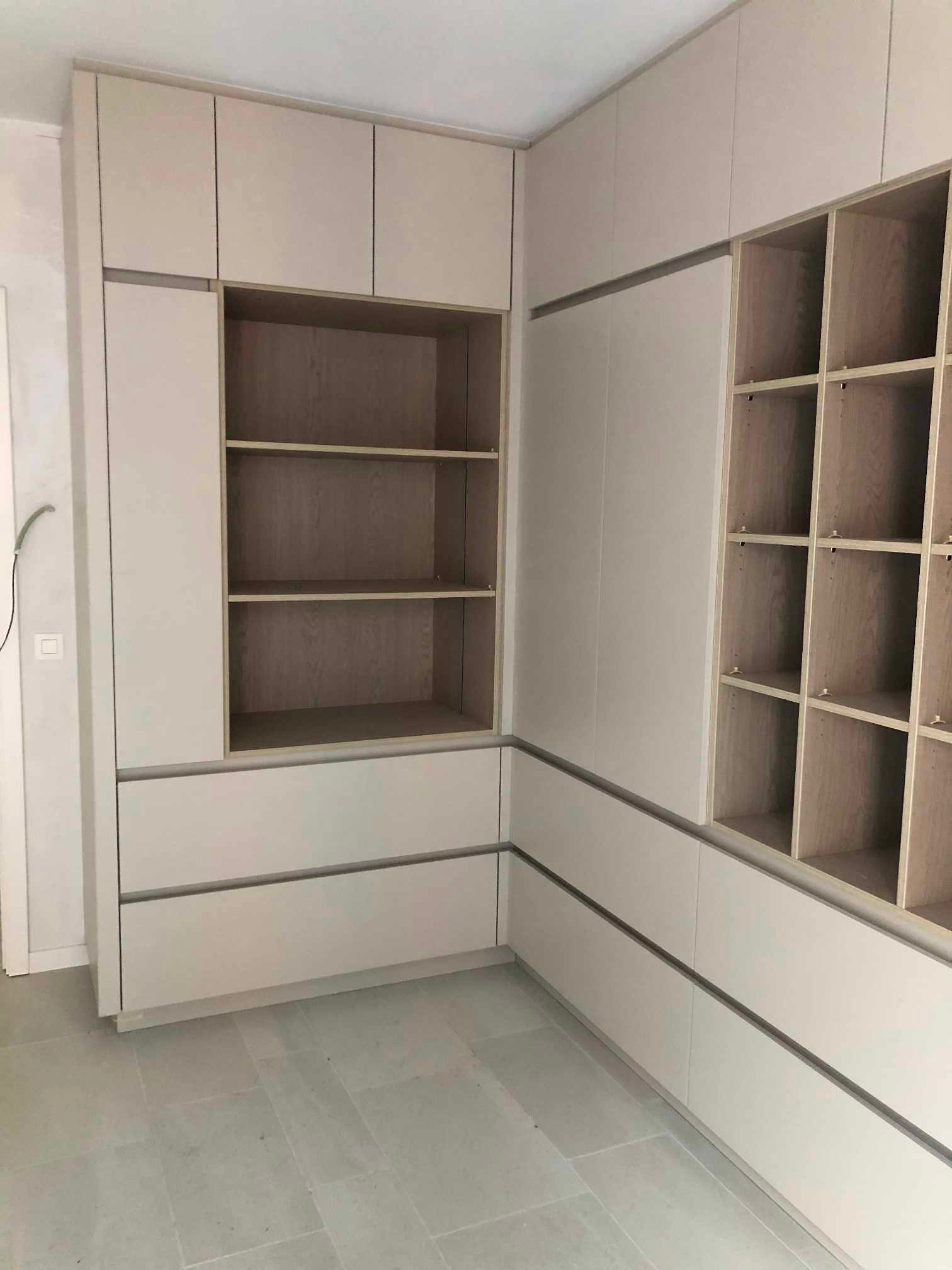 Jasna zabudowa meblowa z szufladami i otwartymi półkami z surowego drewna, widok z rogu pomieszczenia na szarej podłodze.