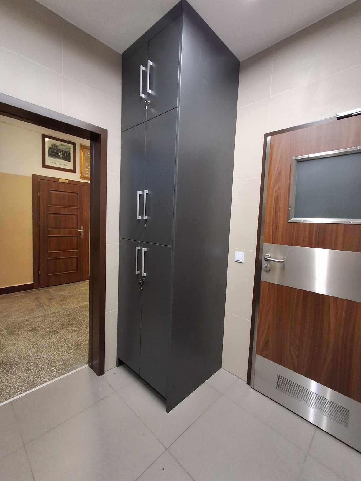 Wysoka, grafitowa szafa z czterema drzwiami i srebrnymi uchwytami, umieszczona w korytarzu obok drewnianych drzwi z metalowymi akcentami i małym okienkiem.